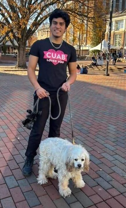 Grinning student holding the leash of a small-medium sized white dog. Both are looking at the camera, and the student's shirt reads: "CUAB loves you."