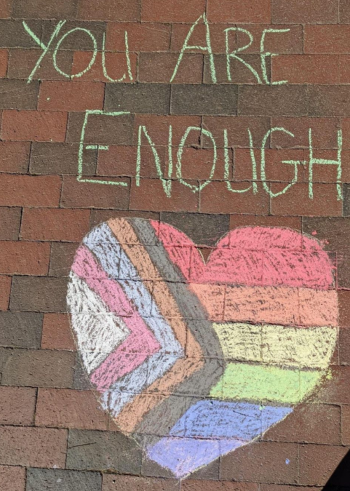 A brick path with a beautiful chalked heart made up of the rainbow flag and an indentation of the colors of the transgender flag and black and brown stripes to represent people of color, and the words "YOU ARE ENOUGH"