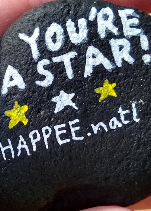 Black painted rock that says in white letters, "You're a star!", with white and yellow stars and @HAPPEE.natl