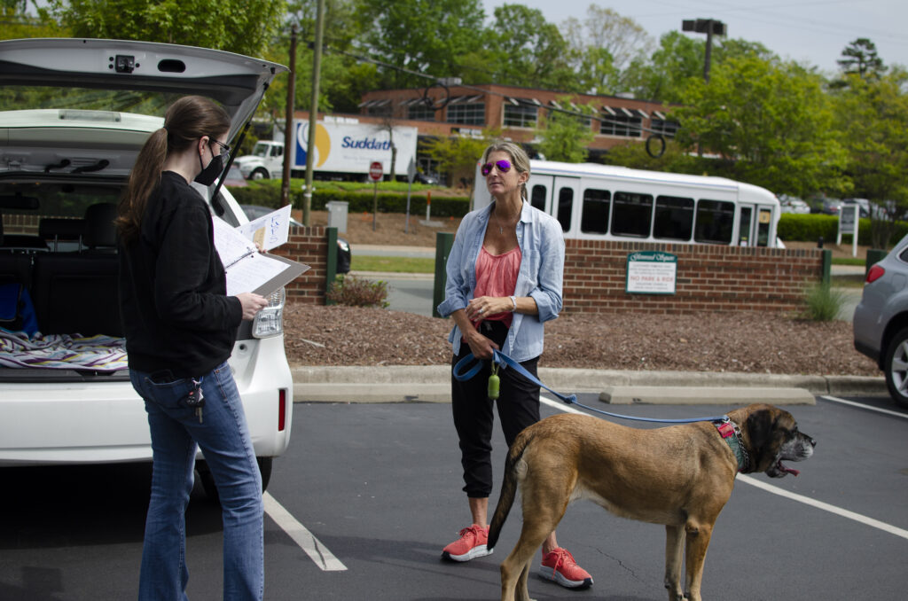 Two women in face masks, talking in a parking lot. One has notes in her hand, the other is holding the leash of a beautiful brown dog (Macie the Mastiff)