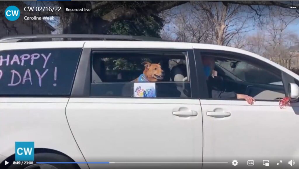 Thumbnail for Carolina Week feature on HAPPEE. A white van is driving by, with "Happy Heart Day" written on the back window, a fairly large brown dog's head (hence his name, Big Dog) with his head sticking out of the middle window smiling, above a sign with the HAPPEE logo. In the front passenger's seat is a man in a face mask, waving to the camera.