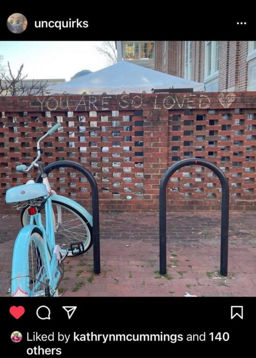 A screenshot of an IG post by UNCQuirks, showing a place to park bicycles with one Carolina blue bike, in front of a half brick wall. At the top of the wall is written, "You are SO loved!" in chalk.