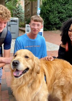 3 students squatting and grinning at Dusty the Golden Retriever