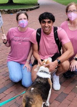 Two masked volunteers on either side of a grinning student who is petting Bobo. All three humans are wearing pink shirts.