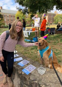 A student smiling and petting Daisy the Golden Retriever who is sitting on the grass wearing a cone-shaped birthday hat. Both are smiling at the camera.