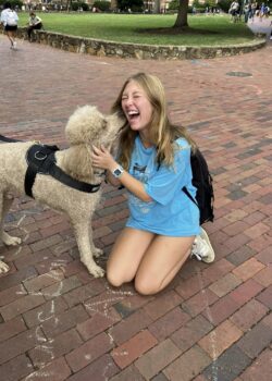 A kneeling student laughing with delight as Apollo, a tan poodle, licks her cheek