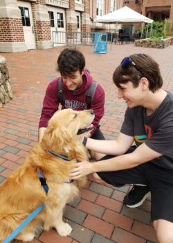 Two students squatting by Dusty the golden retriever, grinning and petting him