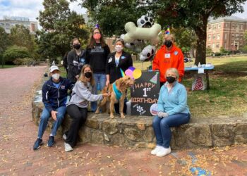 A group of non-student volunteers (and one student volunteer) and Daisy the Golden Retriever around a sign that says "HAPPEE 1st Birthday" and in front of a bone-shaped helium balloon