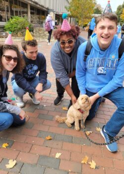 Four students wearing cone-shaped birthday hats around Otis the Cavapoo