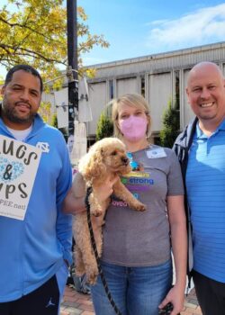 Sean May holding a "Hugs & Pups" sign, Otis the Cavapoo being held by him Mom, and Pat Sullivan standing together